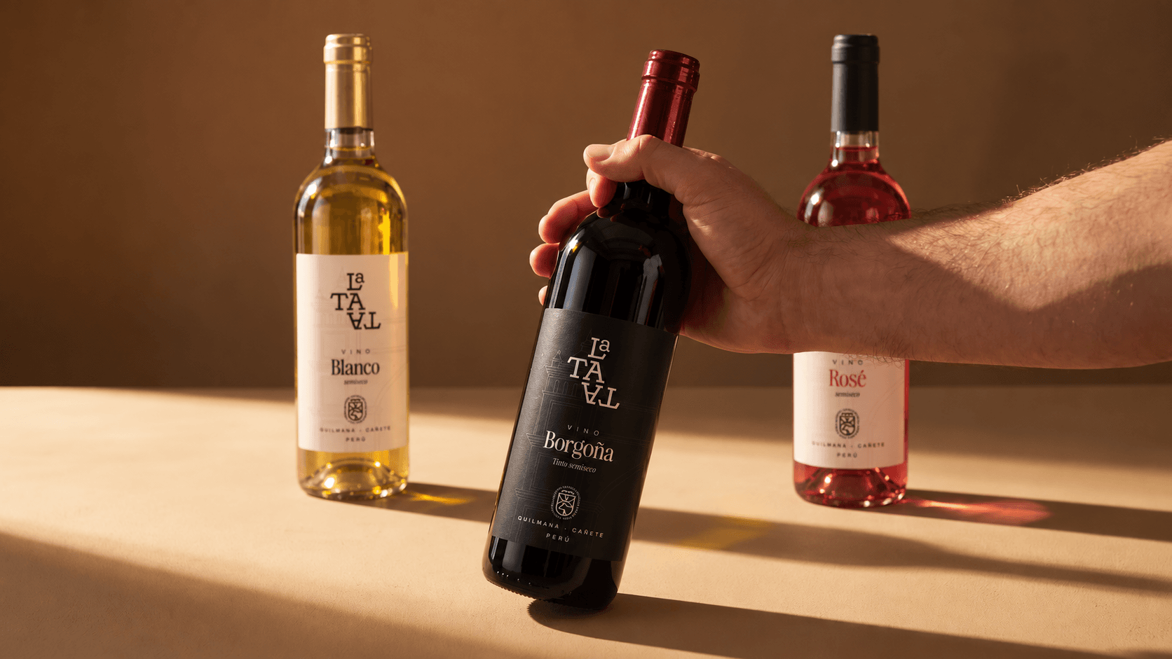 Una mano sostiene el vino Borgoña La Tata junto a las botellas de Blanco y Rosé